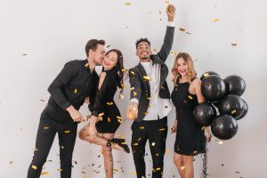 Handsome man kissing his girlfriend standing on one leg in light room. Excited african guy in white shirt throwing confetti on blonde woman with party balloons.