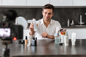 Smiling young man filming his video blog episode about new cosmetic products while sitting at the kitchen table and applying make-up